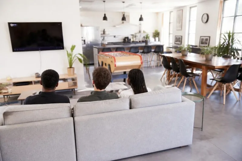 Espace commun d'une résidence avec canapés, télévision, baby-foot et cuisine partagée, trois jeunes vus de dos